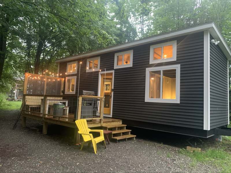 Tiny house on wheels at dusk with warm glowing windows, string lights on the deck, and a yellow Adirondack chair
