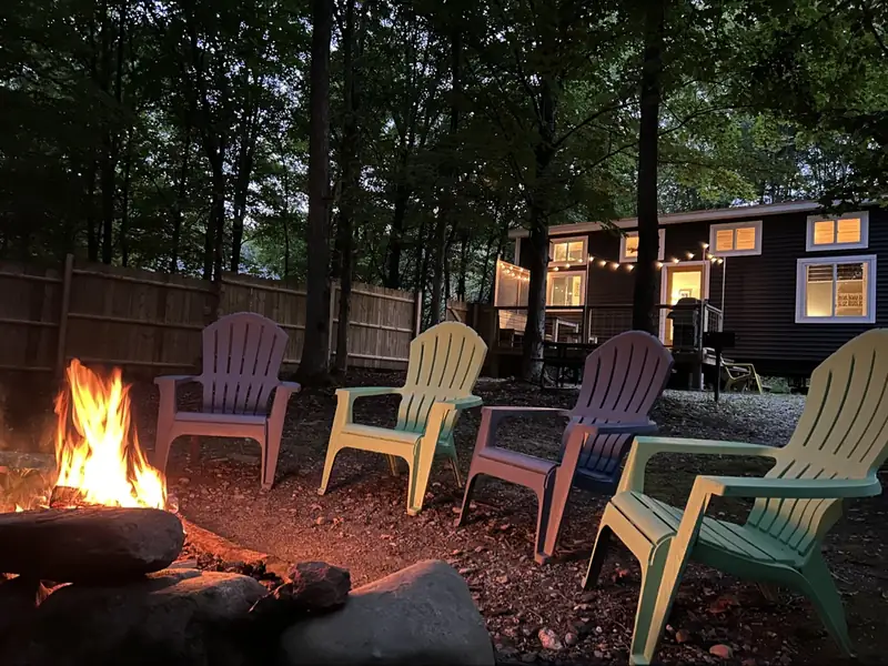 Fire pit burning at night with colorful Adirondack chairs and the glowing tiny house in the background