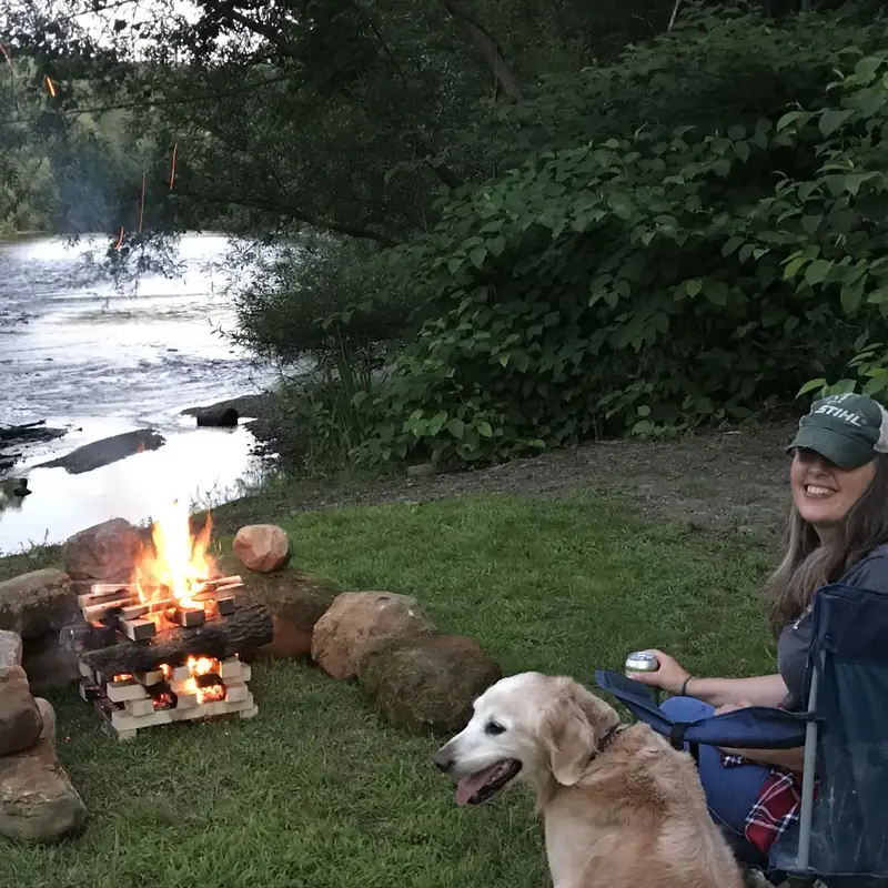 Evening at the riverside fire pit — relaxing by the fire with a dog as sparks rise, the Battenkill River just behind