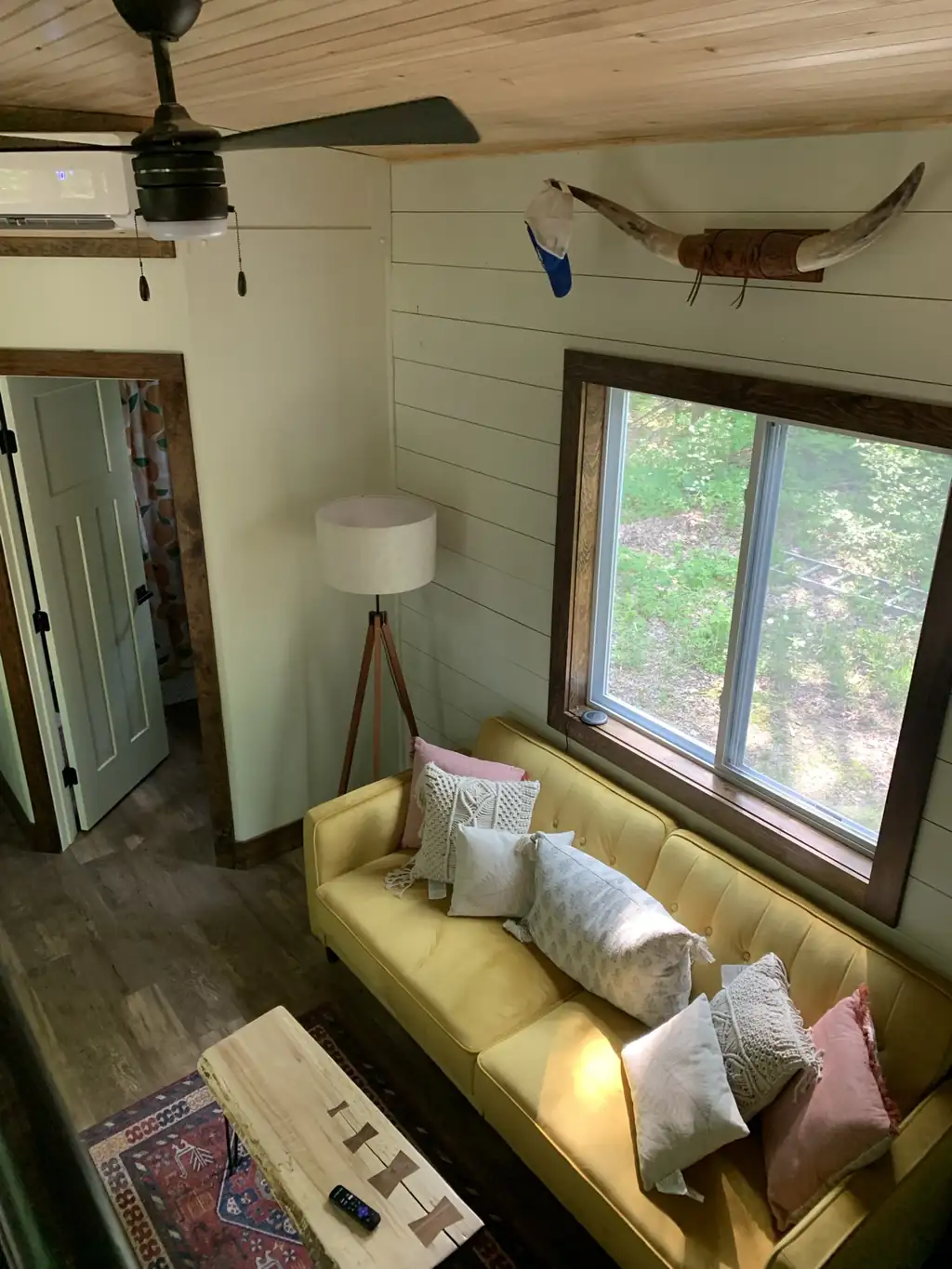 Tiny house living room in daylight: mustard-yellow couch under a longhorn mount on the shiplap wall, with a ceiling fan
